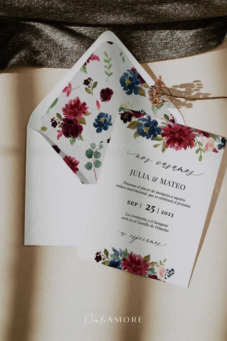 Invitación de boda con flores acuareladas en tonos vino, rosa y azul, acompañada de sobre blanco forrado con ilustraciones florales, sobre fondo neutro con luz suave.