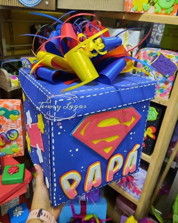 Caja de regalo azul con decoración de Superman y la palabra "Papá", con moño multicolor en la parte superior.