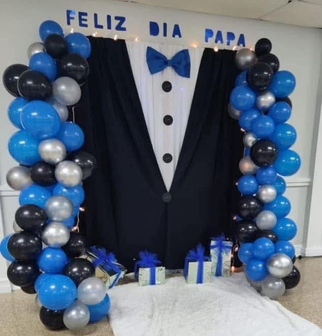 Decoración de fondo con forma de traje y moño azul, acompañado de columnas de globos azules, negros y plateados con el mensaje "Feliz Día Papá".