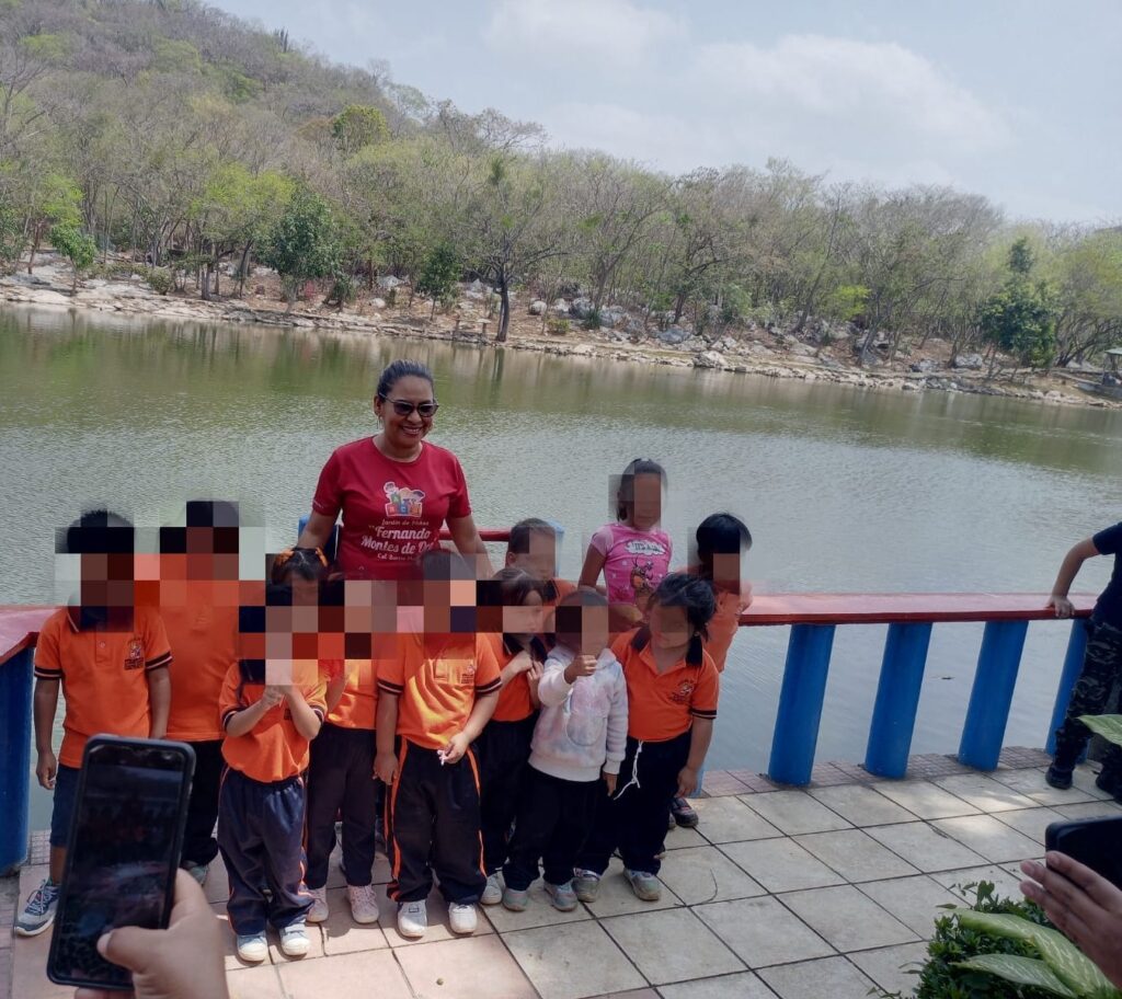 Grupo de niños en uniforme naranja junto a su maestra durante excursión a una laguna en Juchitán