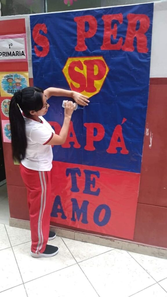 Niña decorando un cartel grande con el mensaje “Súper Papá te amo”, en colores rojo, azul y amarillo.