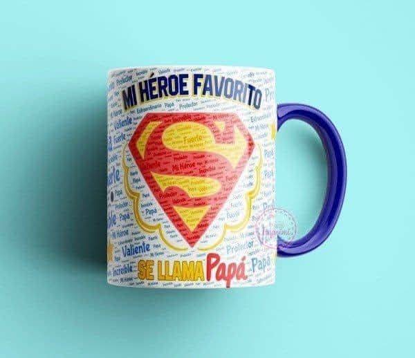 Taza blanca con asa azul, ilustrada con el logo de Superman y frases como “Mi héroe favorito se llama papá”.
